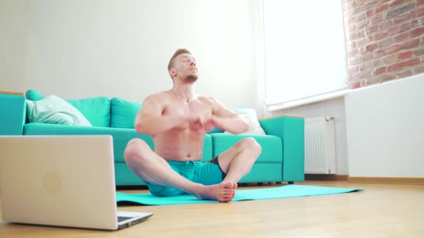年轻的健美男运动员在客厅里用笔记本电脑在地板上练习网上瑜伽 男人赤身裸体做运动 拉长身体距离或拉枕头 录像辅导 — 图库视频影像