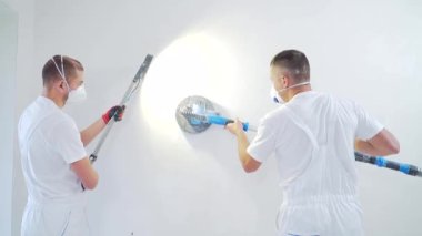 Duvar kağıdı zımpara makinesi zımpara makinesiyle sıva düzleştirici duvar yüzeyi. Beyaz alçı bir duvarı zımparalayan iki usta inşaatçı. Tulumlu bir adam yüzeyi solunum cihazıyla öğütüyor. deneyimli tamirci