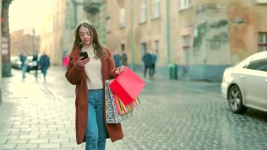 Paltolu genç bir kadın, elinde telefonla sokakta satış yaptıktan sonra alışverişe çıkıyor. Hediye çantalı mutlu çekici kız şehir arka planında şehirde dolaşıyor. Noel hediyeleri