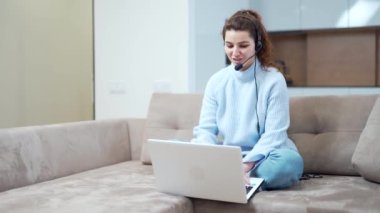 Çevrimiçi mesafe öğrenimi. Evde çalışan kadın internet üzerinden iletişim kuruyor. Öğretmen uzaktan izah eder. Kanepede oturan genç bir kız öğrenci. Kulaklığı ve bilgisayarı var ve webcam ile konuşuyor..