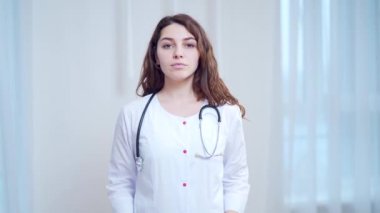 Portreyi kapat. Hastanede duran, beyaz doktor önlüğü ve steteskop iğnesi giymiş gülümseyen genç bir kadın doktor. Mutlu kadın terapist, doktor, kameraya bakan genel pratisyen.