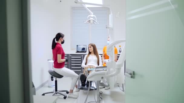 若い女性歯科医は 治療や現代の歯科クリニックでの手順を開始する前に 患者に話します 秘書の専門医がオフィスで女性の相談について説明します — ストック動画