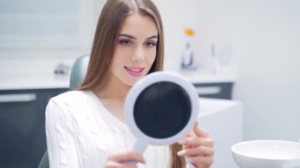 在牙科诊所里迷人的成年女病人看着镜子里的牙齿 快乐而微笑的年轻女性坐在椅子上 在美白或治疗后检查牙齿 — 图库视频影像