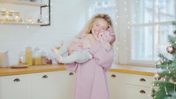 幸せな母は自宅の台所で彼女の小さな娘と遊ぶ ママ赤ちゃんを持ち上げて 暖かいニットの服の抱擁とキスで若い女性幼児の女の子のクリスマスツリーをカダリングボンディングを楽しむ — ストック動画