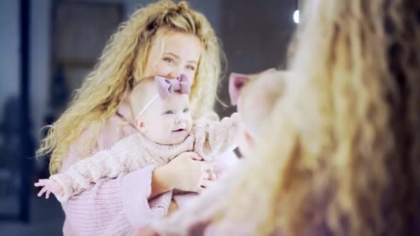 年轻快乐的母亲在家里的镜子前和婴儿玩耍 妈妈们穿着一件针织毛衣 和一个头戴蝴蝶结的女婴一起微笑在他怀里 母亲节 — 图库视频影像