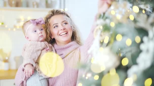 6ヶ月の小さなかわいい母親の肖像画を閉じる古い女の子は自宅でクリスマスツリーを飾る ニットセーターのブロンドのお母さんと赤ちゃんは お正月を祝います Blurred Bokeh — ストック動画