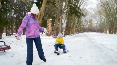 Genç anne parkta karda küçük bir çocukla birlikte parkta yürür. Mutlu anne oğluyla kayıyor. Birlikte vakit geçirin. Soğuk mevsimde çocuklarla dışarıda vakit geçirmek. Açık havada.