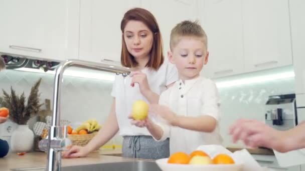 在现代的白色厨房里 有小孩的年轻家庭 两个小男孩帮助他们的父母做饭 把水果洗干净 一起呆在家里 爸爸和妈妈在家里有礼貌的孩子们 — 图库视频影像