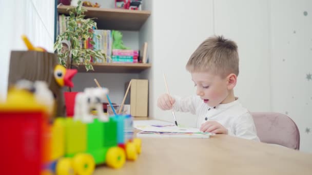 一个小男孩用画笔和彩色颜料在桌边作画的特写 在家里休闲的孩子有一种最喜欢的创造性爱好 快乐的金发小孩画画 — 图库视频影像