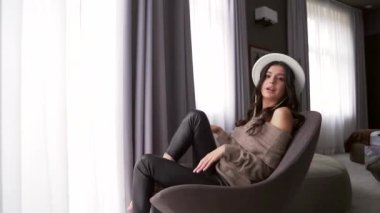 Çekici ve seksi bir kadının portresi. Bir sandalyede otururken otel ya da apartmanda bacak bacak bacak üstüne atmış. Çekici moda kadını, şapkalı manken kameraya baştan çıkarıcı bir şekilde bakıyor..