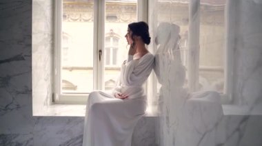 Sabahları sabahları sabahlık giymiş zarif bir gelin. Lüks banyo iç çamaşırlı, beyaz iç çamaşırlı çekici bir kadın pencerenin yanında pencere kenarında oturuyor. Baştan çıkarıcı bir şekilde kameraya bakıyor..