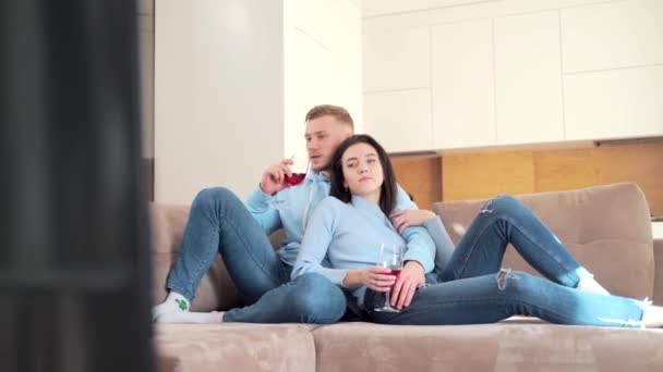陽気な若いカップル ボーイフレンドとガールフレンドのテレビを見てソファに座ってワインを飲む 家族の男と女は一緒に新しいアパートで自宅でテレビを見て楽しむ余暇を過ごす — ストック動画