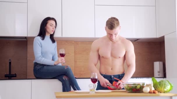 性感的没穿衬衫的小伙子在厨房里做饭 他的女朋友在喝酒 年轻夫妇恋爱中 一起准备早餐或午餐 男人和女人烹调蔬菜沙拉 — 图库视频影像