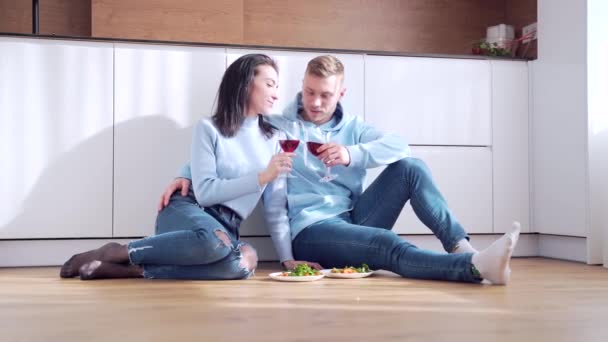 赤いワイングラスでキッチンの暖かい床でリラックスして座っている愛の幸せな新婚夫婦は 新しい改装された家でデートを楽しみ 話して 夢を共有しています 新しいアパートでお祝いする若い家族 — ストック動画