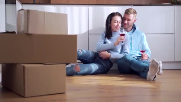若い幸せな夫婦と女性が床に座って新しいアパートや家に移動を祝う 彼らはワインを飲み 段ボール箱の近くのインテリアを計画します 現代的なデザインの夢 — ストック動画