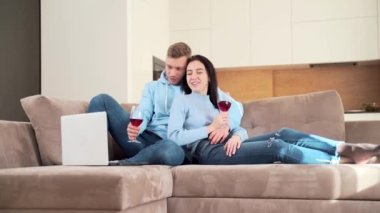 Genç çift kanepede dinleniyor, şarap içiyor ve dizüstü bilgisayara bakıyor. İnternete bak ve konuş. Sohbetiniz kutlu olsun. Oturma odasında vakit geçirin.