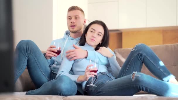 陽気な若いカップル ボーイフレンドとガールフレンドのテレビを見てソファに座ってワインを飲む 家族の男と女は一緒に新しいアパートで自宅でテレビを見て楽しむ余暇を過ごす — ストック動画