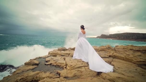 在大海或大海的背景下 穿着白色婚纱和面纱站在岩石上的年轻新娘 在风景秀丽的背景下 迎面而来的海浪吸引着自然界中迷人的年轻女子 这让人惊叹 — 图库视频影像