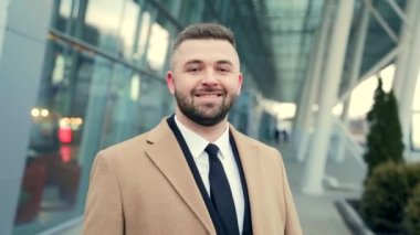 Kapalı portre genç yakışıklı iş sakallı adam kentsel arka planda gülümsüyor modern bina kameraya bakıyor. Resmi takım elbiseli ve ceketli neşeli bir iş adamının yüzü.