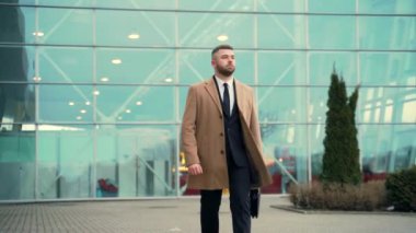 Genç, şık, sakallı bir iş adamı elinde ceket ve çantayla şehir caddesinde yürüyor. Modern bir binanın kentsel arka planında. Başarılı ve kendine güvenen bir iş adamı portresi