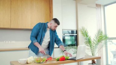 Genç yakışıklı sakallı adam evde dizüstü bilgisayarla yemek hazırlıyor. Erkek, vejetaryen salatası için sebze keser ve yemek tarifini öğretici video dersleri izler.