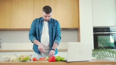 Genç yakışıklı sakallı adam evde dizüstü bilgisayarla yemek hazırlıyor. Erkek, vejetaryen salatası için sebze keser ve yemek tarifini öğretici video dersleri izler.