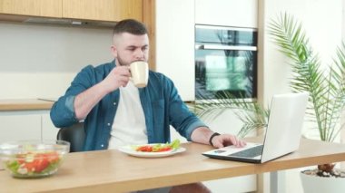 Genç, yakışıklı, sakallı, evde çalışan ve mutfaktaki masada yemek yiyen serbest bir erkek. Kahvaltı ya da öğle yemeği. Yemek masasında meşgul bir adam dizüstü bilgisayarla sabahları evde kahve içiyor.