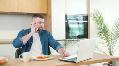 Genç, yakışıklı, sakallı, evde çalışan ve mutfaktaki masada yemek yiyen serbest bir erkek. Kahvaltı ya da öğle yemeği. Yemek masasında meşgul bir adam dizüstü bilgisayarla sabahları evde kahve içiyor.