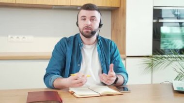 Kulaklık toplantısı, konferans ya da iş kursu, giysi ya da video bağlantısı olan sıradan giyinmiş genç sakallı bir adam. Adam kamerayla konuşuyor. Webcam görüntüsünde çalışıyor. Açıkla ve uzaktan konuş.