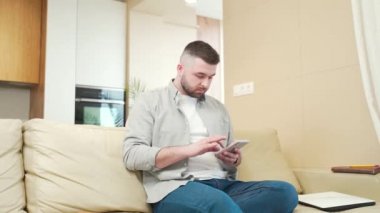 Cep telefonlu heyecanlı genç adam evdeki kanepede oturmuş spor bahsini kazanıyor, iyi haber, telefonuyla açık artırmayı kutlayan neşeli adam açık artırmayı kazanan oyunu kazanıyor, SMS alıyor.