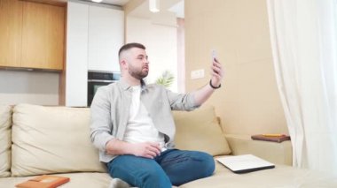 Günlük giysiler içinde neşeli genç sakallı adam konuşuyor ve iletişim kuruyor. İnternette cep telefonuyla ön kameraya gelen bir görüntüyü kullanarak, evdeki bir kanepede oturarak.