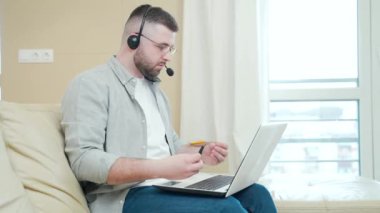 Genç sakallı iş adamı internette video konferansında çalışırken evinde kulaklığı olan bir dizüstü bilgisayar kullanıyor. Erkek işadamı web kamerası eğitim ağı pazarlamasını uzaktan açıklıyor