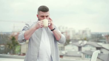 Modern balkonda ya da terasta dizüstü bilgisayarla uzaktan çalışan genç, yakışıklı sakallı adam şehir manzaralı iş adamı, açık hava ofislerinde serbest giyiniyor.