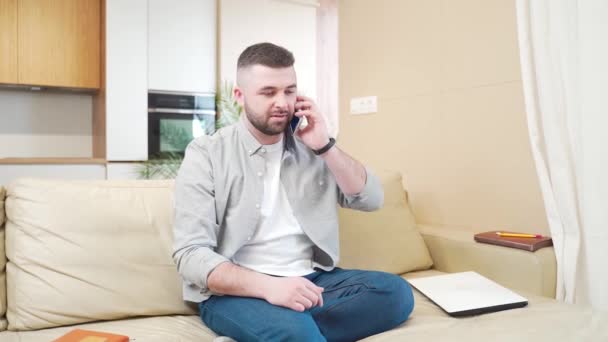 身穿休闲装 留着胡子的年轻人说话和交流 在客厅里坐在沙发上的手机上 男性讲智能手机 — 图库视频影像