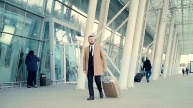 Takım elbiseli ve ceketli genç sakallı erkek turist havaalanından bir valizle ya gelir ya da gider. Modern bir binanın arka planında. Şık bir iş adamı şehir gezisi