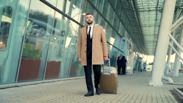 ビジネススーツとコートを着た若いひげを生やした男性観光客は スーツケースの到着または出発で空港から行きます 近代的な建物の裏側にあります スタイリッシュなビジネスマンの街の旅を歩く — ストック動画