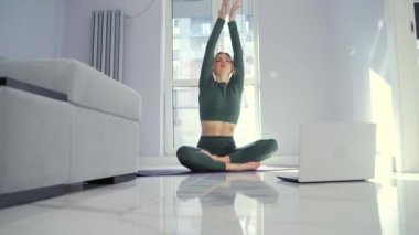 Spor kıyafetli genç sarışın kadın video dersleri kullanarak internette yoga yapıyor. Evde, oturma odasında, dizüstü bilgisayarın önünde, yerde oturan spor salonundaki kadın. Siluet içeri yansıtılıyor