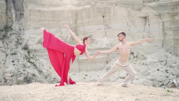 若い情熱的なプロのダンサーのカップルが自然の上の山の上で屋外で一緒に踊ります 岩や砂の背景に赤いドレスを着た男と女 ロマンチックなモダンバレエ — ストック動画