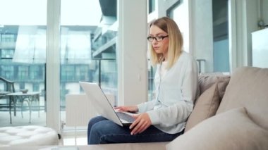 Genç bir iş kadını internette çalışıyor bir kanepede dizüstü bilgisayarla oturuyor ve webcam 'de sohbet ediyor. Sarışın bir iş kadını video görüşmesi yapıyor. Konferans veya sohbet mesafesi