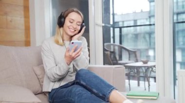 Evdeki kanepede oturup kulaklıkla müzik dinleyen neşeli, genç bir kadın. Mutluluk Yaşam Tarzı. Genç kız ya da öğrenci hayattan zevk alır. Elinde cep telefonuyla gülümsüyor. Uygulama kullanılıyor