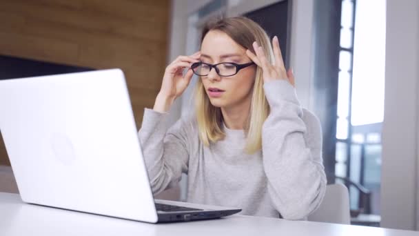 年轻的金发女人戴着眼镜在笔记本电脑上工作 头疼得很厉害 在工作或家里的电脑里的女学生很疲倦 痛死了女人的头 — 图库视频影像
