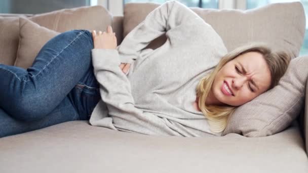 年轻妇女坐在或躺在沙发上或沙发上 腹部剧痛 女性在床上用经前 经期或中毒来控制她的胃 金发女孩腹部抽筋 — 图库视频影像