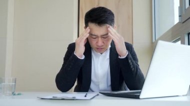 Genç Asyalı bir adam, ofis çalışanı iş yerinde oturuyor el ele tutuşuyor alnına ve kafasına şiddetli bir acı ile masaj yapıyor. Baş ağrısı olan bir erkek. Izdırap çeken kişinin yüzüne aşırı derecede hastalık bulaşmış. Kapalı