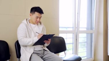 Genç Asyalı öğrenci bekleme odasında bir sandalyeye oturup başvuru formu dolduruyor. Erkek hasta resepsiyonda ya da iş yerinde ya da teste girmeden önce. Hasta koridorda bekleniyor.
