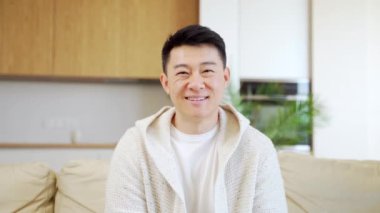 Kapalı koltukta otururken kameraya bakıp gülümseyen Asyalı bir genç portresi. Evdeki kanepede günlük giysiler içinde mutlu bir erkek öğrenci. Salondaki tatmin olmuş adam. Kapat.