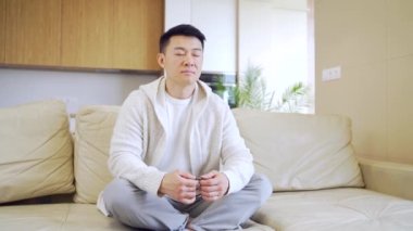 Sakin, düşünceli Asyalı genç bir adam kanepe evinde gözleri kapalı oturur, meditasyon yapar ve yalnız başına dinlenir. Erkek, Lotus pozisyonunda oturup yoga yapıyor. Etrafında barış ve uyumun tadını çıkarıyor. Denge uyumu. Rahatla.