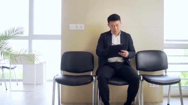 年轻的亚洲男人穿着正装在候车室里等待面试或会面的结果 商界男性在位 学生通过考试前 坐在椅子上等工作 — 图库视频影像