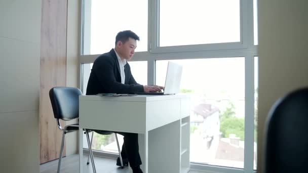 オフィスのパソコンやノートパソコンに座って仕事をしているアジア系の男性のシルエット 街を見下ろす窓の前で残業で一人でオンラインで働く男性マネージャー — ストック動画