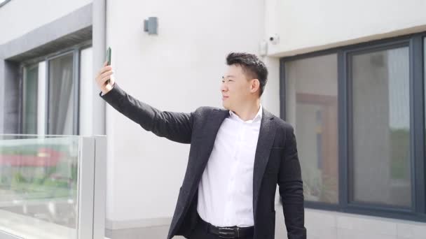 若いアジアのビジネスマンが携帯電話やスマートフォンのカメラを使ってオンラインでビデオ通話をしています 屋外テラスでのビジネス男性は笑顔と手で手を振って正式なスーツで電話 — ストック動画