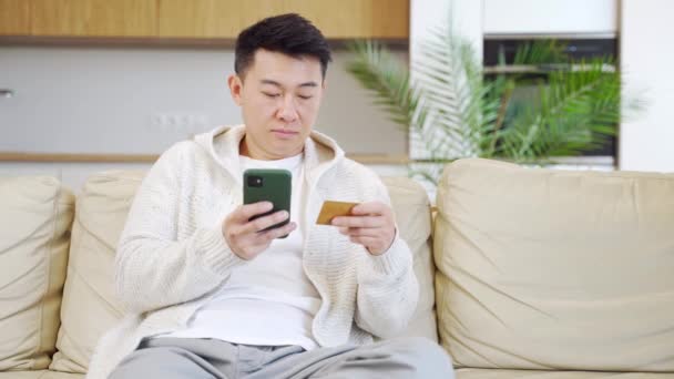 携帯電話やクレジットカードを使ってネットショップで買い物をしている若い幸せなアジア人男性です 男性は自宅のソファに座っているアプリケーションでサービスやチケットの支払いデータを入力します 簡単なアプリ — ストック動画